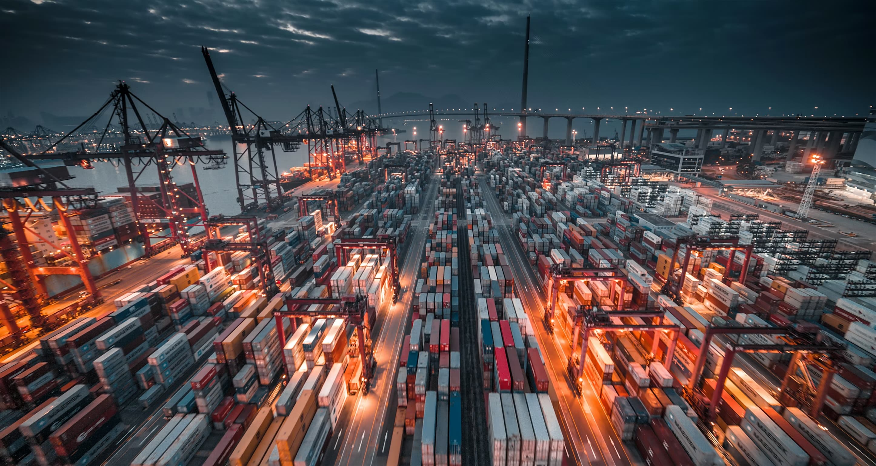 Elevated view of a major container port at dusk: ship-to-shore cranes, stacked terminals and the bridge and city beyond